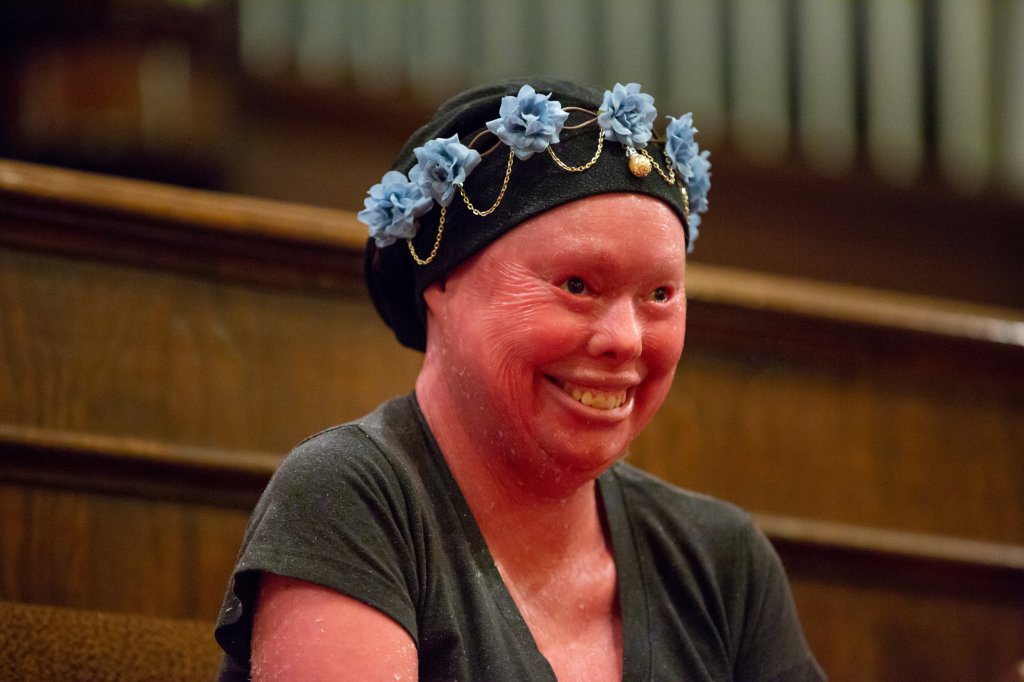 Image is of Hunter Steinits, M.Div. She has red skin and is smiling. She is wearing a grey top and a navy blue hat with light blue flowers and gold chain around the front crown of the hat. 